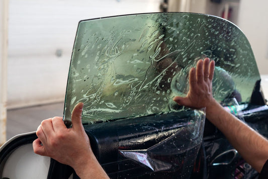 A person applies a tinted film to a car window with soapy water.