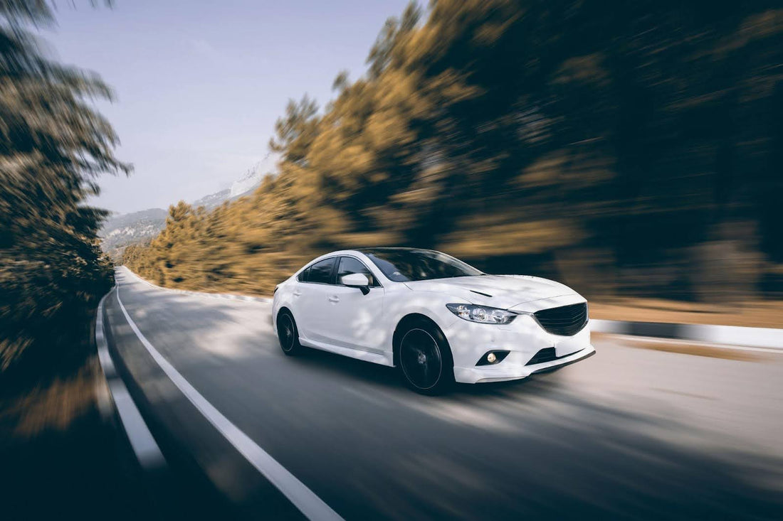 A white Mazda sedan in motion on a road with trees lining the sides