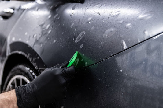 Hand in a black glove using a green sponge to clean a wet car surface.