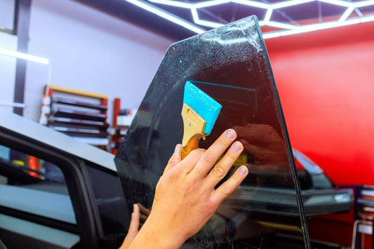 Hand using a blue squeegee to apply a clear film to a car window in a workshop.